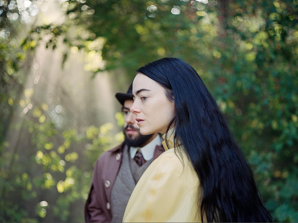 Actor Emma Stone stands in the centre of the frame, facing left. She has light skin and long dark blue hair. She is wearing a light yellow top and standing in a sun-dappled grove of trees. Behind her, actor Ramy Youssef stands facing the left of the screen. He has a beard, moustache and black hat. He is wearing a burgundy jacket over a grey tweed vest and white button-up shirt.