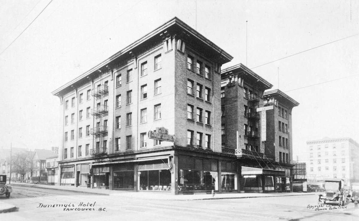 A black and white photo shows the Dunsmuir Hotel in 1923. The photo is taken from an angle that shows the front and eastern side of the hotel. It is a five-storey building with commercial businesses on the bottom level and four floors of hotel rooms above.