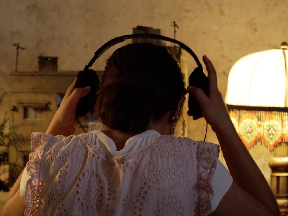 A photograph of the back of a girl putting on a pair of headphones in the living room of a dimly lit home. She is wearing a white cap-sleeved T-shirt under a light pink dress with eyelets. There is a tabletop lamp on the right and a potted fern on the left.