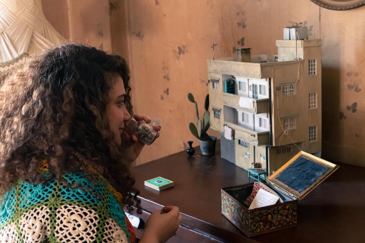 A young woman with shoulder-length brown curly hair is seated to the left of the screen, wrapped in a blue, green and white crocheted quilt. She holds a small glass container to her nose and looks towards the open box and brown cardboard diorama of an apartment on the table before her.