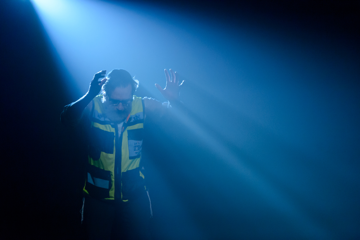 A middle-aged man in glasses, a beard and a neon yellow safety vest stands to the left of the screen in a ray of blue light against a darkened background. He is looking down with both hands raised, elbows bent, above his shoulders.