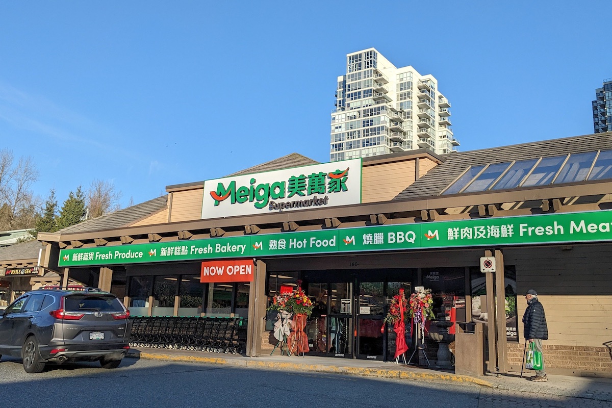A supermarket with green branding in a suburban plaza. There are signs in English and Chinese. A tower rises from the back.