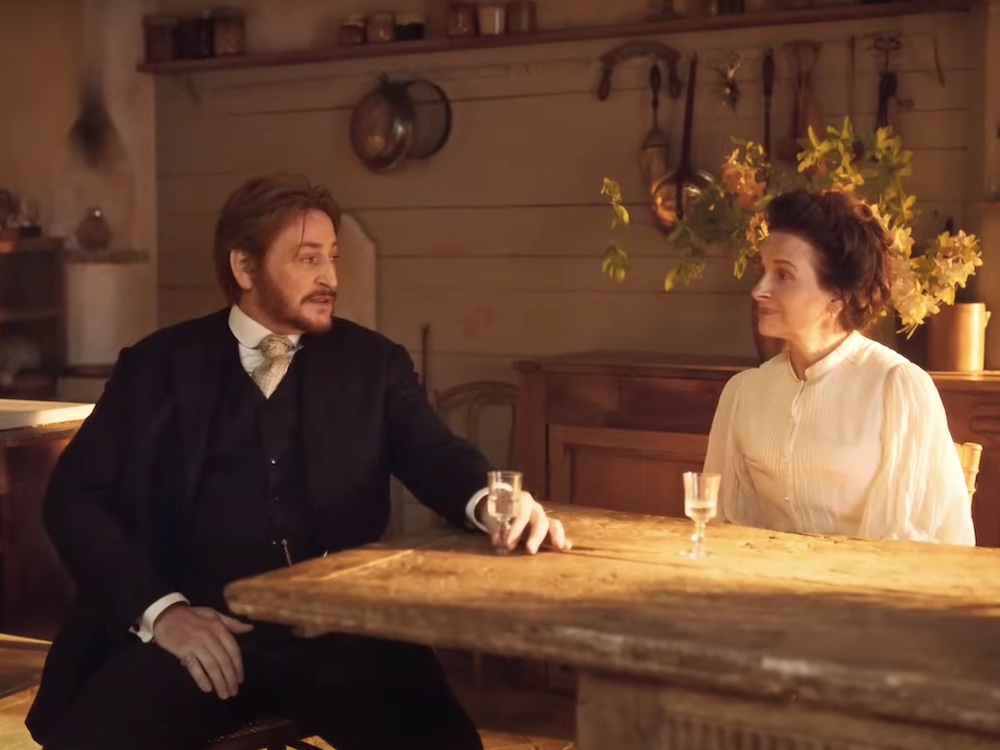 A man and a woman with brown hair and light skin sit at a long wooden kitchen table in golden light, looking at each other. They are wearing formal wear from the late 1800s. The man on the left is in a black suit. He holds the stem of a small white wineglass at the table. The woman on the right has her hair loosely up. She is wearing a white dress and looking at the man. Behind her is a yellow bunch of flowers in a vase on a wood cabinet.