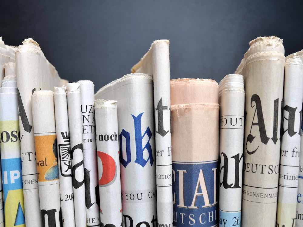 A stack of folded newspapers stands vertically, pressed tightly together, as though they are books on a shelf. They are against a blue background.