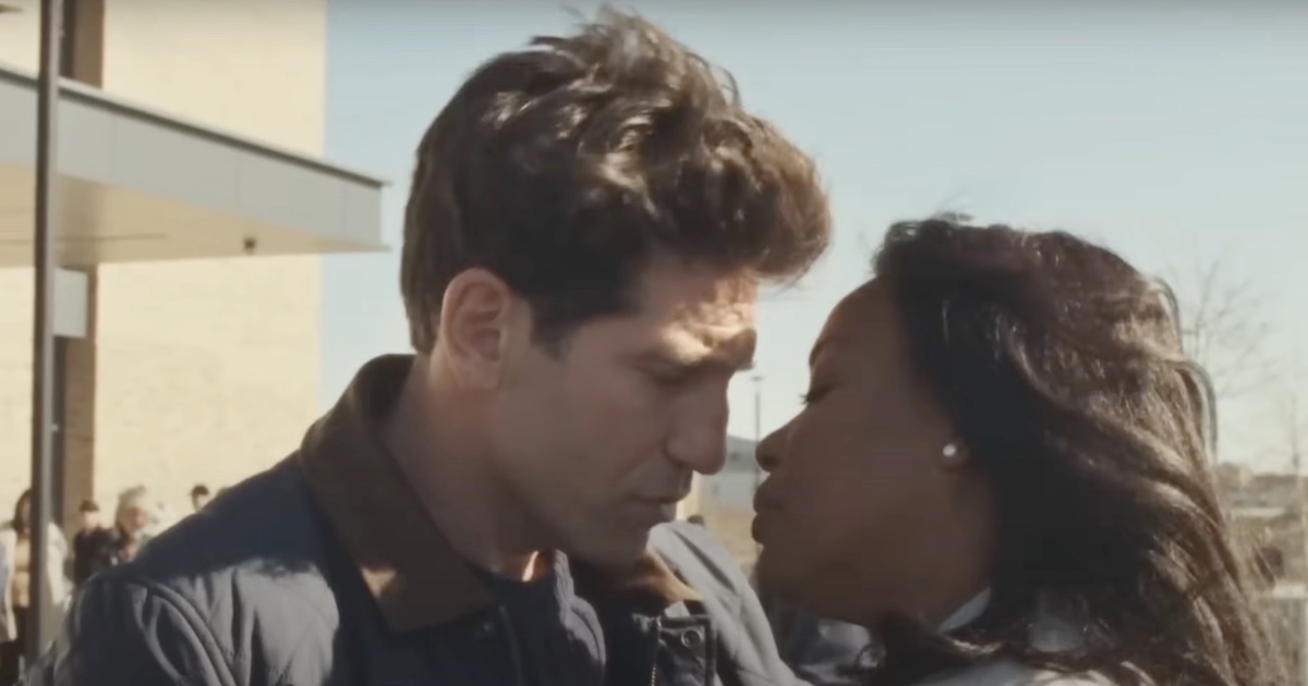 A white man, left, leans towards a Black woman, right, as they move to kiss. They are standing outdoors in a sunny space with a blue sky in the background.