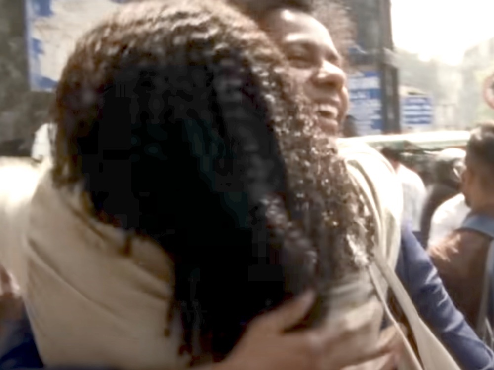 Two Black people embrace each other on a busy downtown street on a sunny day, with one person’s back turned to the camera. The person who is facing the camera is looking up, smiling brightly.