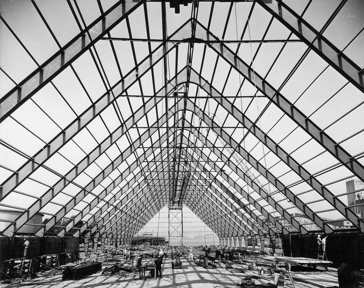 A black and white photo from 1956 shows a large triangular space with a roof composed of windows. It is the raw sugar warehouse. Workers can be seen doing daily tasks.