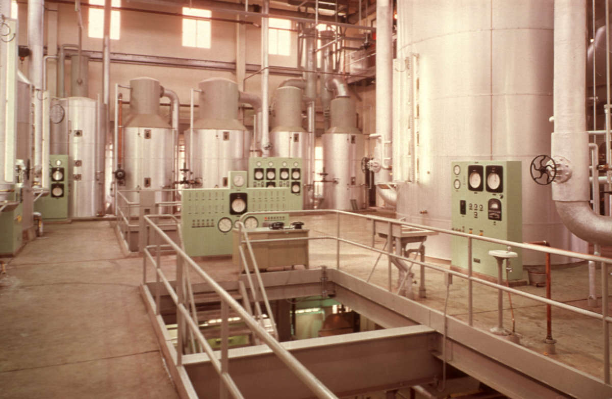 An older photo, from the 1970s, showing the interior of the BC Sugar Refinery. There are a lot of big metal vats that are wider at the bottom and thinner at the top.