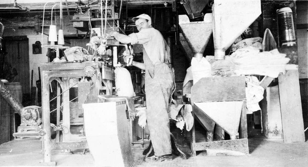 A black and white photo taken in the early 1900s shows a worker in coveralls packing sugar into bags.