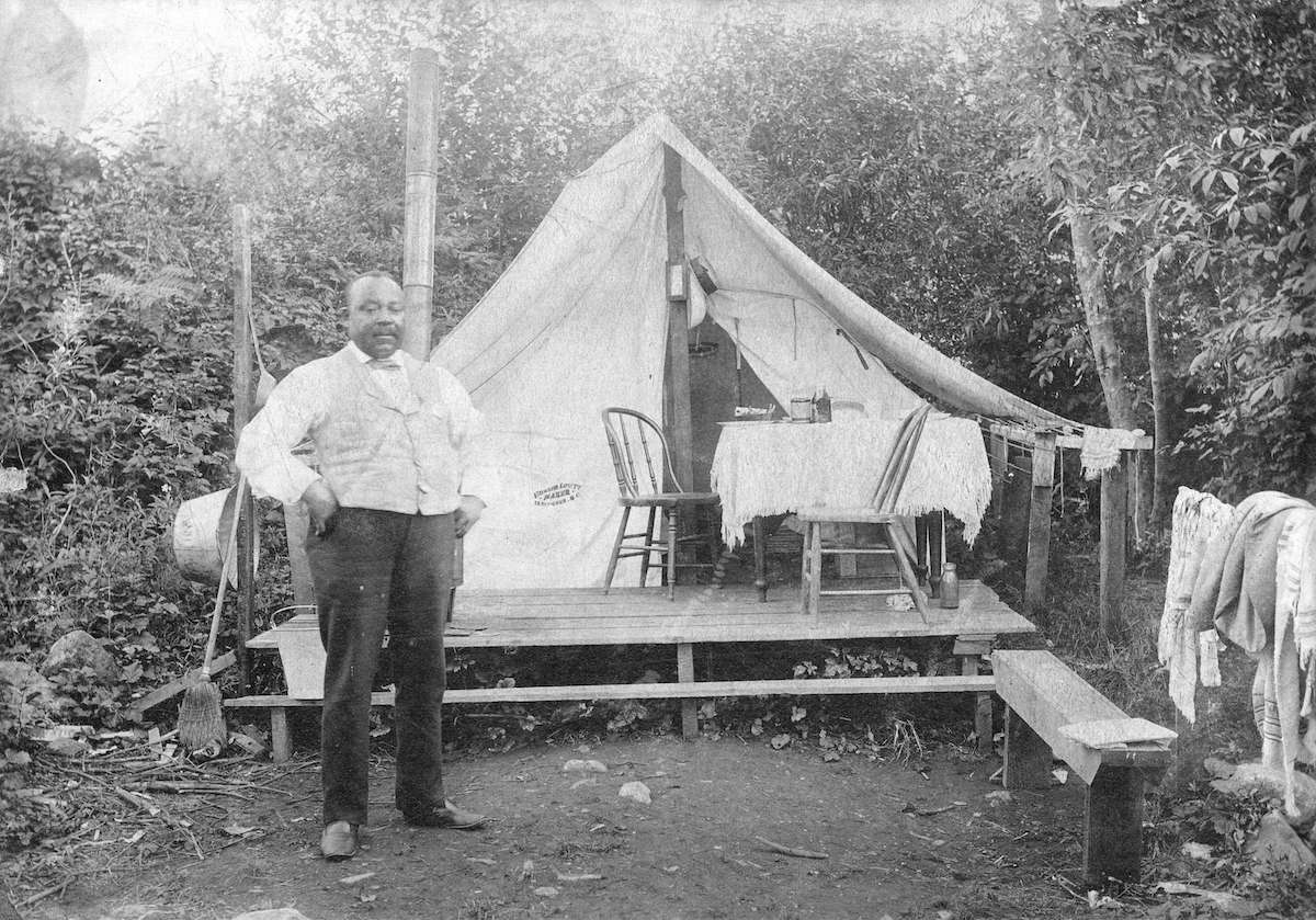 An archival black and white photograph of Seraphim Joseph Fortes features a Black man standing to the left of the frame in a white button-down shirt, a vest, trousers and leather shoes. His hands are on his hips and he is standing in front of a white tent on an elevated wood platform. A pair of wooden chairs are arranged around a table with a tablecloth. The tent is in a clearing against a stand of deciduous trees.