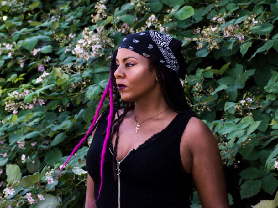 Ruby Smith Diaz is a Black woman with long black hair and bright fuchsia highlights. She wears a navy kerchief in her hair and a black sleeveless top. She stands against a sprawling green blackberry bush with white blossoms. She looks serenely towards the left of the frame.