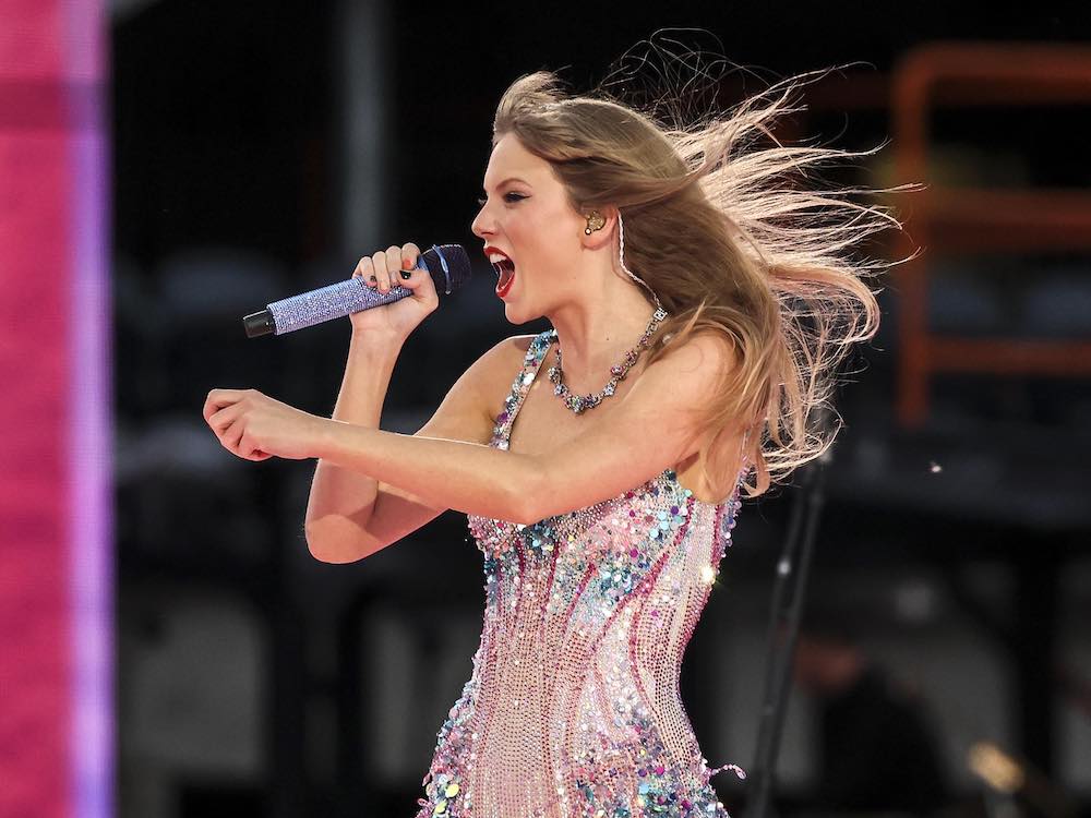Taylor Swift sings into a sparkly silver microphone on an outdoor stage. She is facing the left of the screen and her face is in profile. Her long blond hair blows behind her dramatically. She is wearing a pink sequined bodysuit and bright red lipstick.