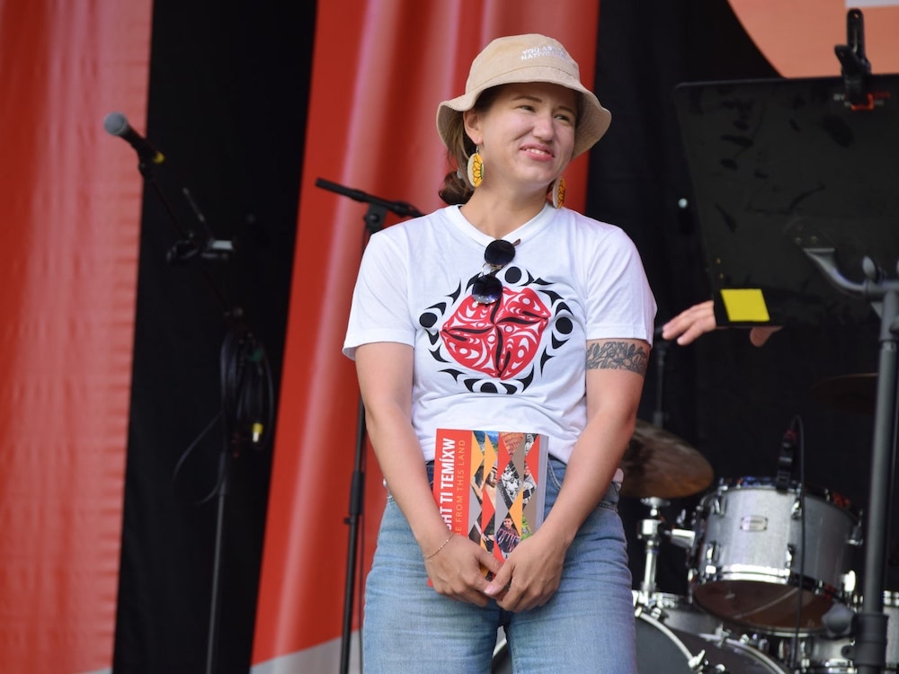 A smiling woman in a beige bucket hat stands on a stage holding a colourful book.