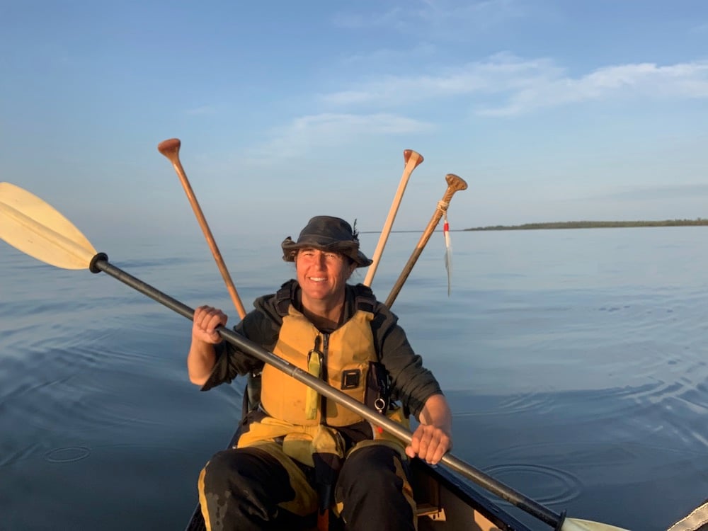 A woman in a yellow lifejacket, black hat and black top paddles towards the camera in a canoe. Behind her is an expansive blue sky and a wide stretch of water on a sunny day.