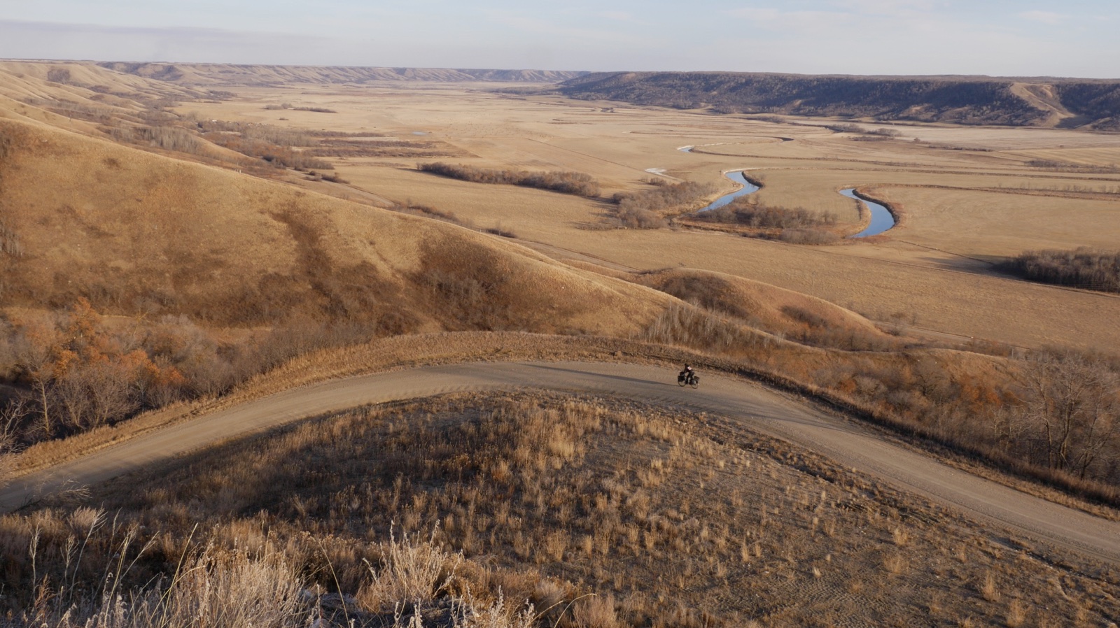 A panoramic photo of rolling golden hills features a distant view of a cyclist biking along an empty, winding pathway.