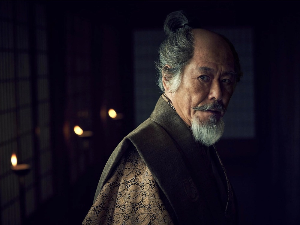 A Japanese man in a historical costume looks over his left shoulder. He has medium-length greying hair tied in a topknot. He has a manicured moustache and beard.