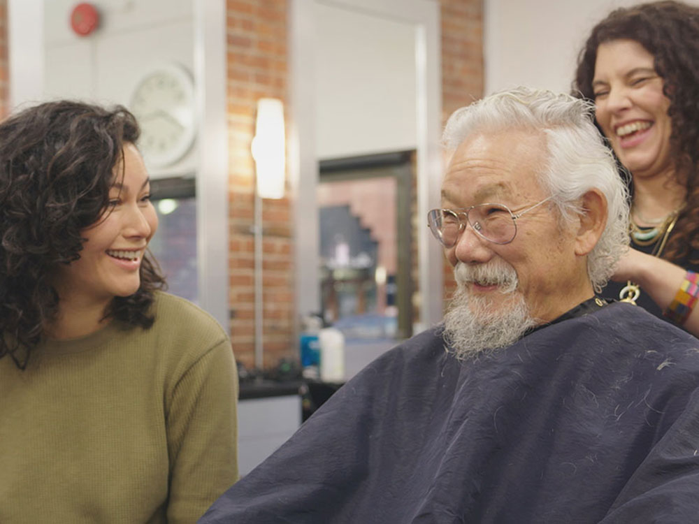 A woman in a green sweater with dark shoulder-length curly hair sits to the left of the frame, looking at her father, a man with white hair, glasses and a beard. He’s smiling and wearing a black barber’s smock. Behind him, the woman cutting his hair is laughing.