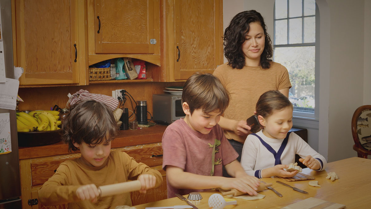 A woman with shoulder-length curly hair and a mustard-yellow T-shirt brushes the hair of a young girl while she cuts out cookies on a kitchen table. To her left are two other children. Behind them is a golden-brown set of cabinetry.