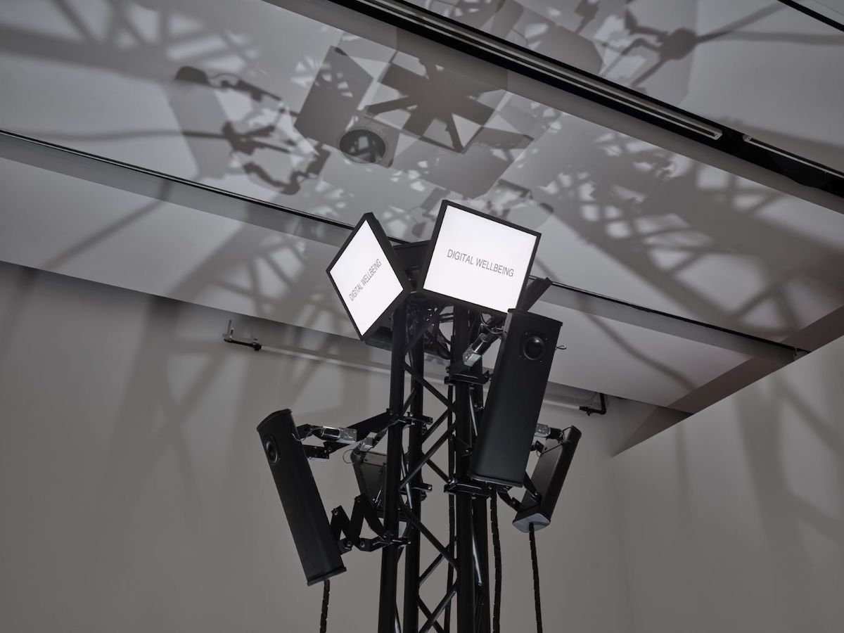 An assembly of digital equipment in the style of surveillance cameras stands atop a thin piece of black scaffolding. Two screens project two words each against white backgrounds: 'DIGITAL WELLBEING.'