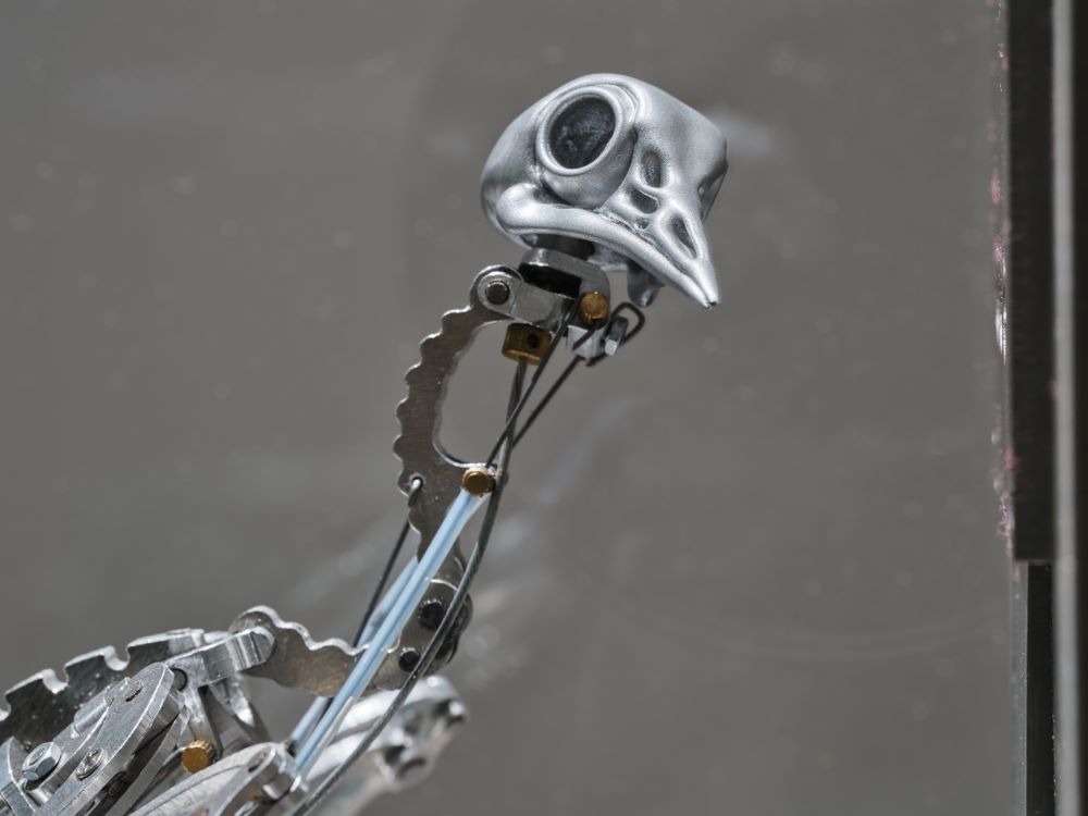 A close-up photograph of a steel skeletal bird looking quizzically at the camera against a grey background.
