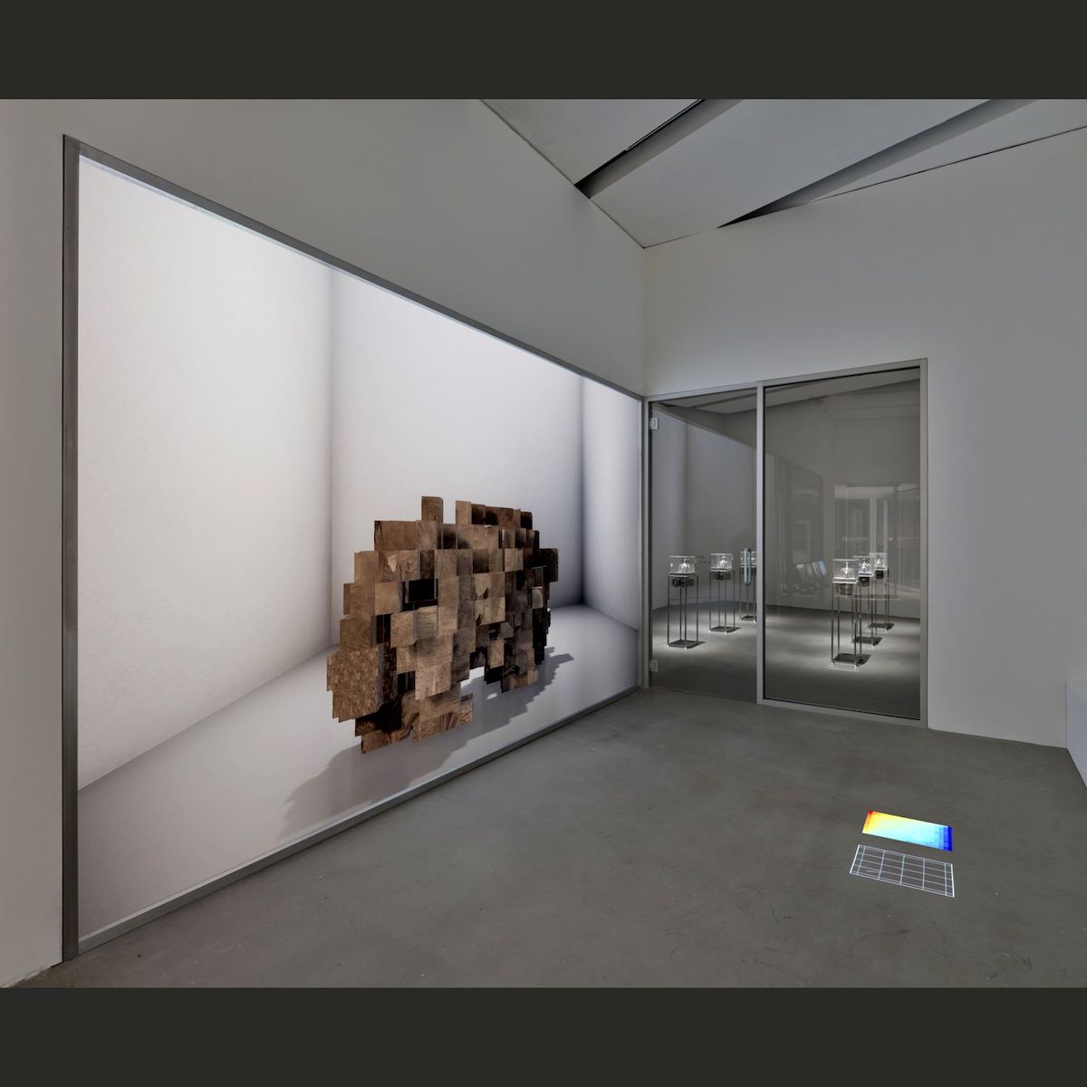 A digital projection of a series of brown pixelated blocks in an indoor gallery space with white walls and grey flooring.