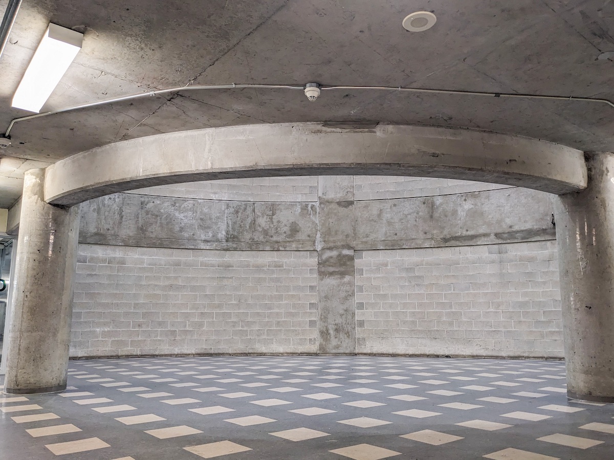 Light illuminates an underground circle that is half open to the parking garage and half bordered by concrete. The floor is a square checkerboard pattern, contrasting with the circular shapes of the lightwell and the columns supporting the lightwell.