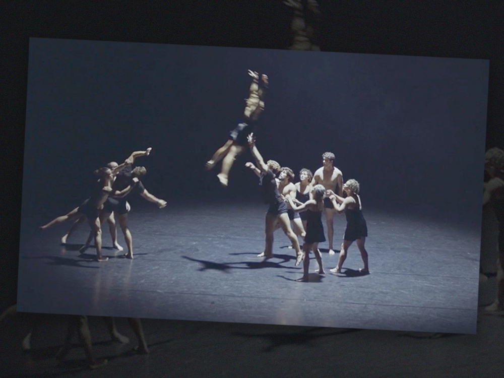 To the right of the frame, a team of dancers in black shorts and short dresses move to catch a dancer falling through the air towards them. They are being thrown from a smaller group of dancers on the left. They are against a black background on a black stage.