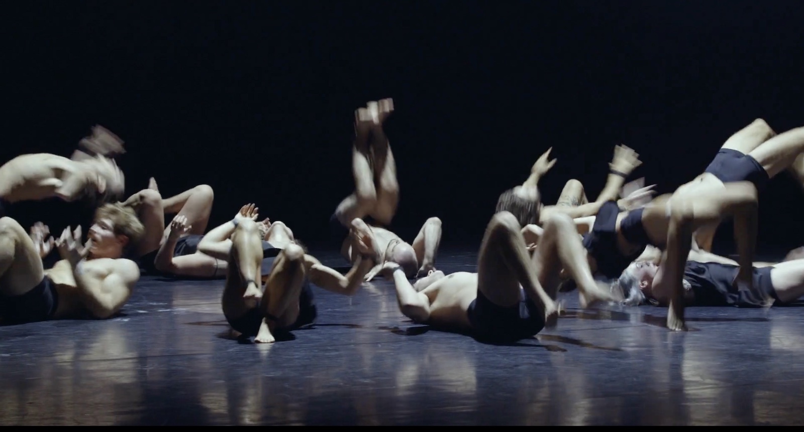 Several dancers are lying on their backs in a circle on a black floor against a black background. They are in the process of moving to rock themselves up on their feet.