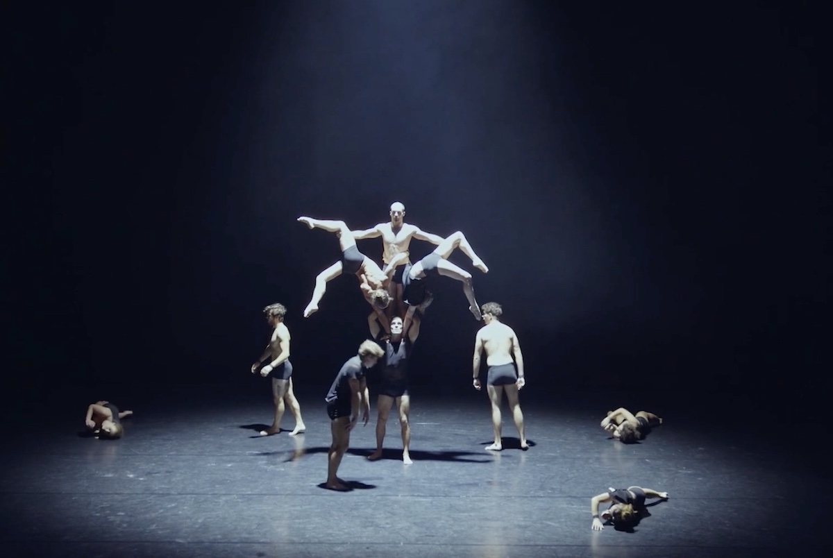 A trio of dancers pose atop one dancer’s shoulders while three others encircle them. The dancers are onstage against a black background.