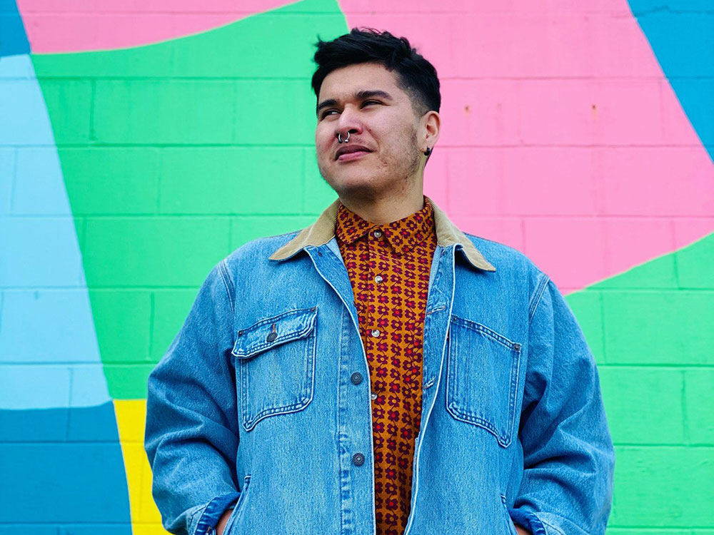 Billy-Ray Belcourt has short dark hair, medium-light skin and a nose piercing. He is looking towards the distance to the left of the frame. He wears a patterned orange button-up shirt under a light blue denim jacket. His hands are in his pockets and he stands against a colourful mural on a brick wall with soft blue, pink, green and yellow shapes.