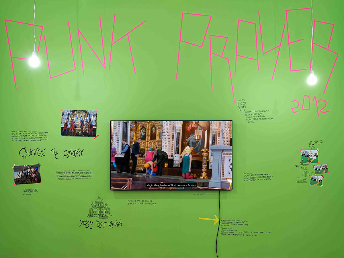 A green wall features hot pink tape forming the letters 'Punk Prayer' around a collection of photographs and a television screen mounted to the wall depicting Pussy Riot performing protest songs at the Cathedral of Christ the Saviour in Moscow.