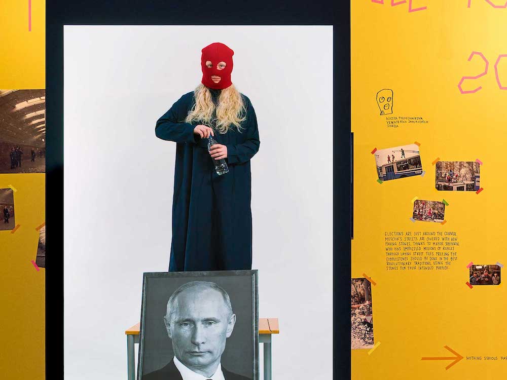 A person with long blond wavy hair and a bright red balaclava stands on a wooden desk against a white background. She is wearing a long black robe and holding a water bottle. A black and white framed portrait of Vladimir Putin leans against the front of the desk below her. In the background are yellow gallery walls decorated with punk ephemera.