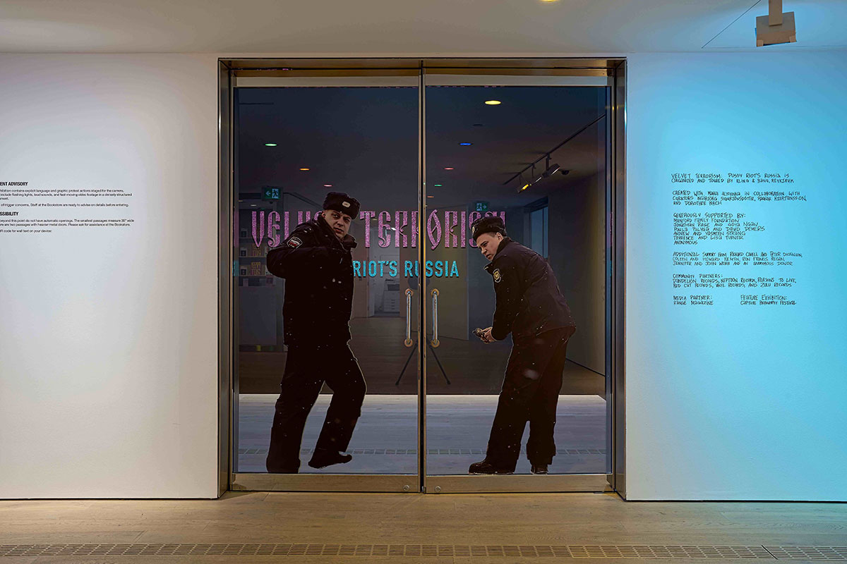 Two security guards stand behind a glass door in dark uniforms and Russian hats as part of an art gallery installation. The walls flanking them are lit blue with exhibition text on them.
