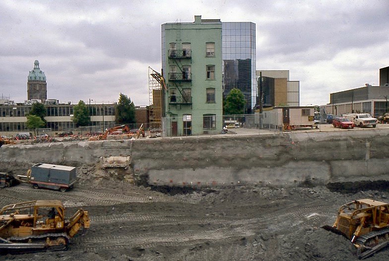 In the foreground, construction equipment rumbles around a large hole in the earth that has been bolstered with concrete walls. In the middle ground, the Del Mar Hotel from the back. It is the only building standing on that side of the street.