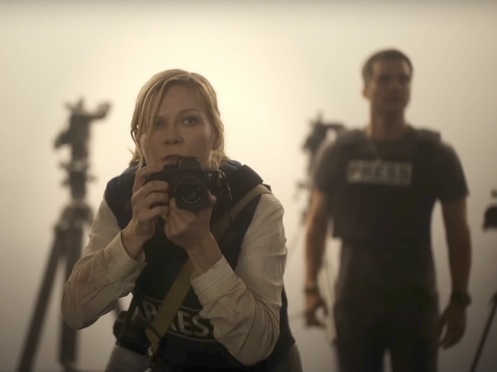 A woman with tied-back blond hair, light skin and pale eyes holds a camera. She wears a white button-down shirt under a dark flak jacket emblazoned with the word 'Press.' Another person with a 'Press' vest stands in soft focus behind her to the right.