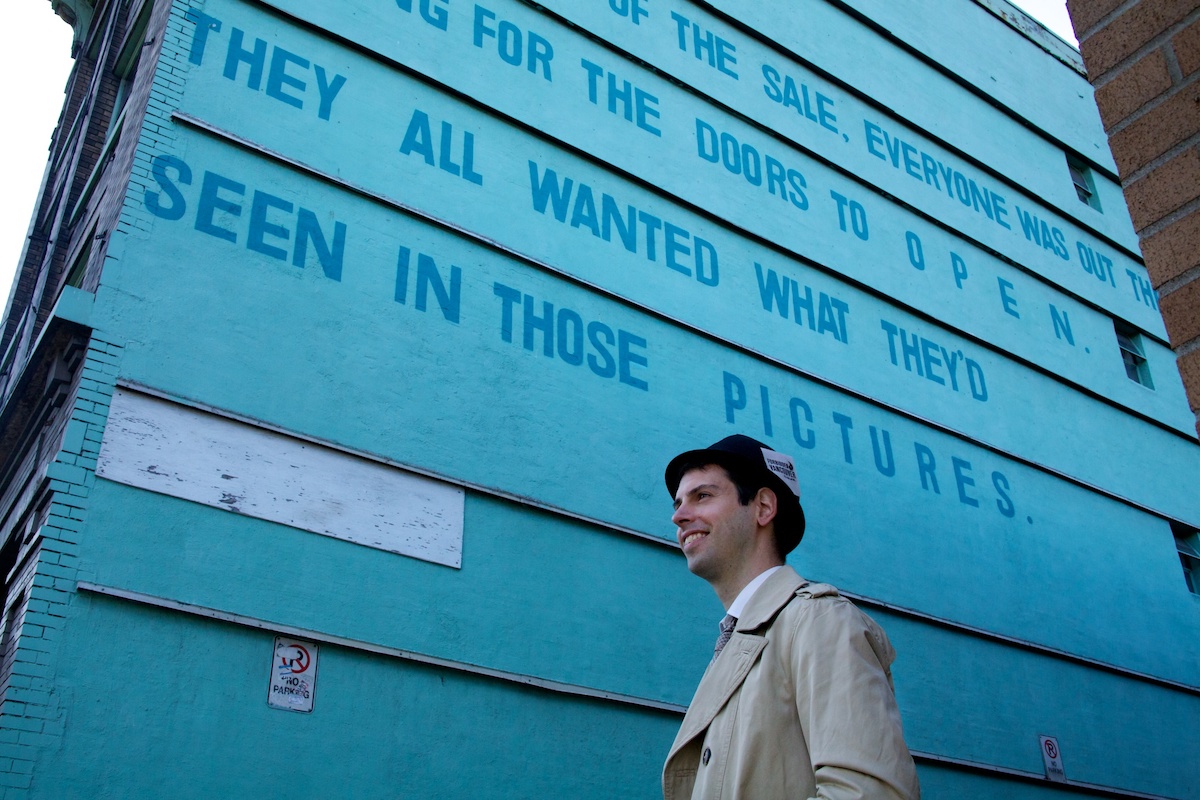 A young man with light skin, wearing a black hat, stands in front of the side of the Del Mar, which has been painted turquoise. The bottom two lines of the mural read, 'They All Wanted What They’d Seen in Those Pictures.'