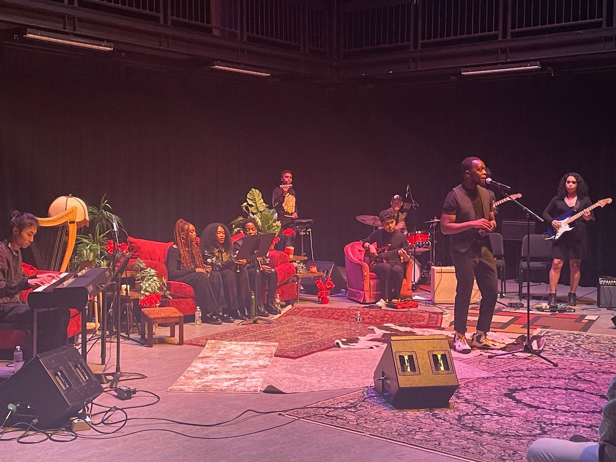 Several musicians in black clothing perform across a stage set up like a living room. Some performers are seated on red sofas and vases of red roses decorate the stage.