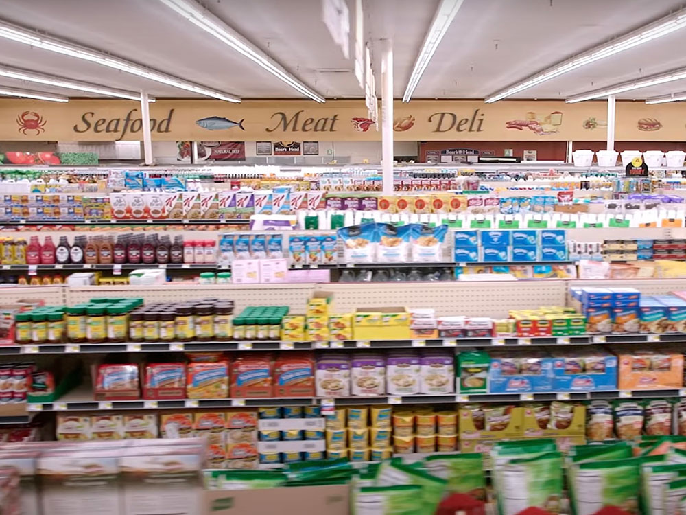 Rows of shelves are stocked with colourful packaged items in a grocery store with low ceilings and fluorescent lighting.