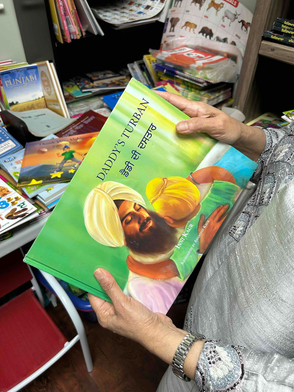 A person wearing a silver suit holds a bright green children&rsquo;s book titled 'Daddy&rsquo;s Turban,' featuring an illustration of a Sikh man wearing a yellow turban embracing a child. In the background is the interior of a bookstore full of books.