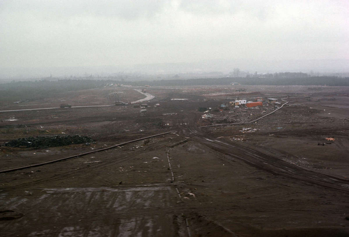 A photograph from 1976 shows a landscape of dirt carved up by piping and roads. There are cars driving across the landscape and a large structure in the upper right section of the image.