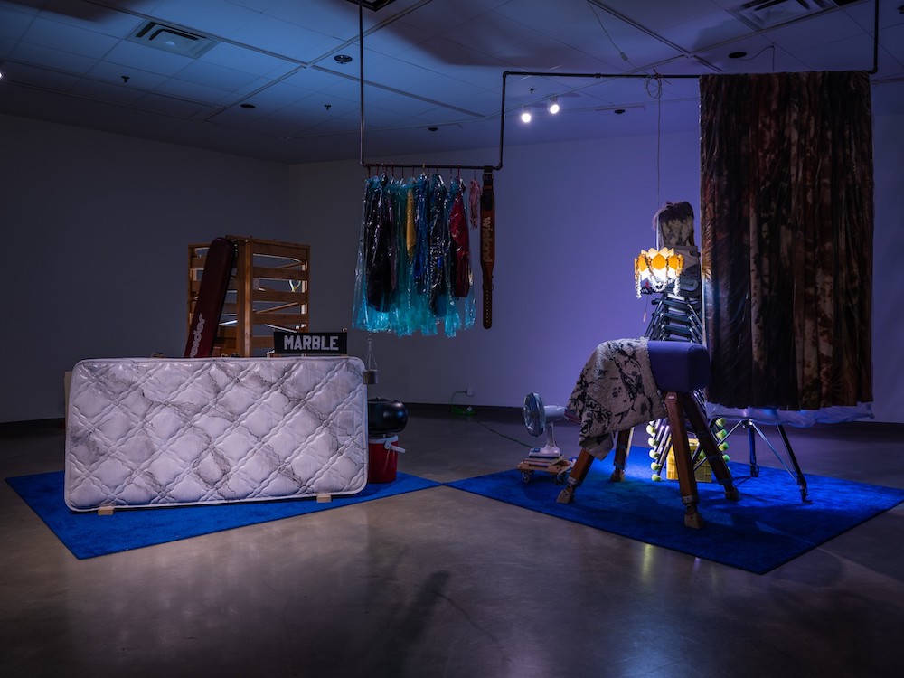 A stage setting evokes a basement, with blue lighting, a mattress standing on its side, clothes in plastic bags on a makeshift rack, a fan and a sign that says 'MARBLE.'