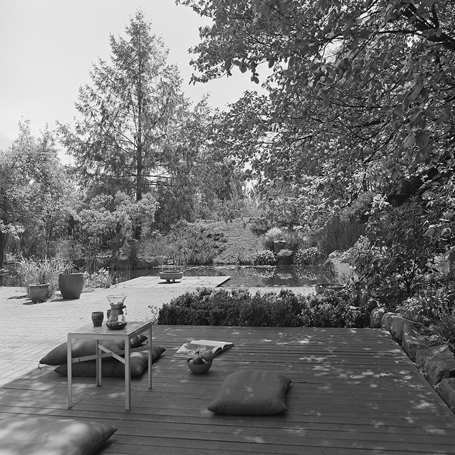 Black and white photo of an elegant back deck with pillow seats. Beyond it is a pond with tall trees behind.