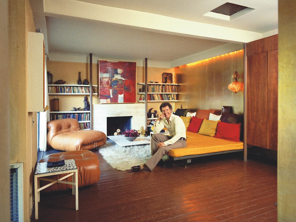 A man sits on a couch smiling in a cozy mid-century home, with warm-toned pillows and fixtures.