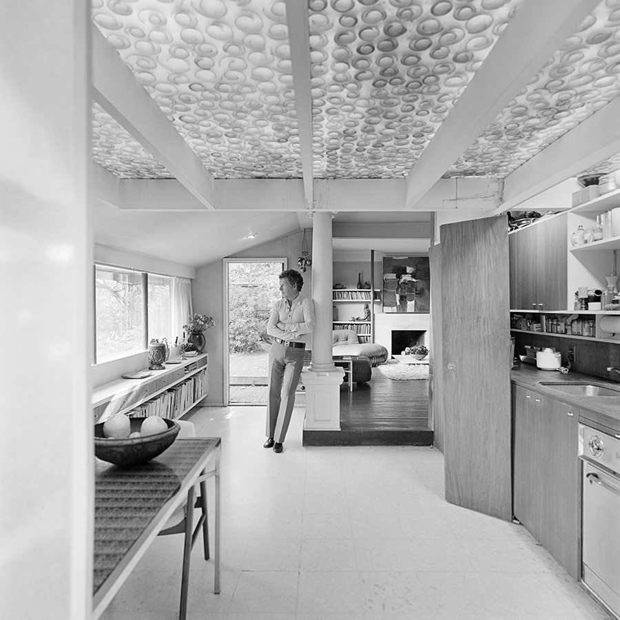 A man leans against a pillar in a 1970s kitchen. The interior of the house is small and cosy, almost like a cottage.
