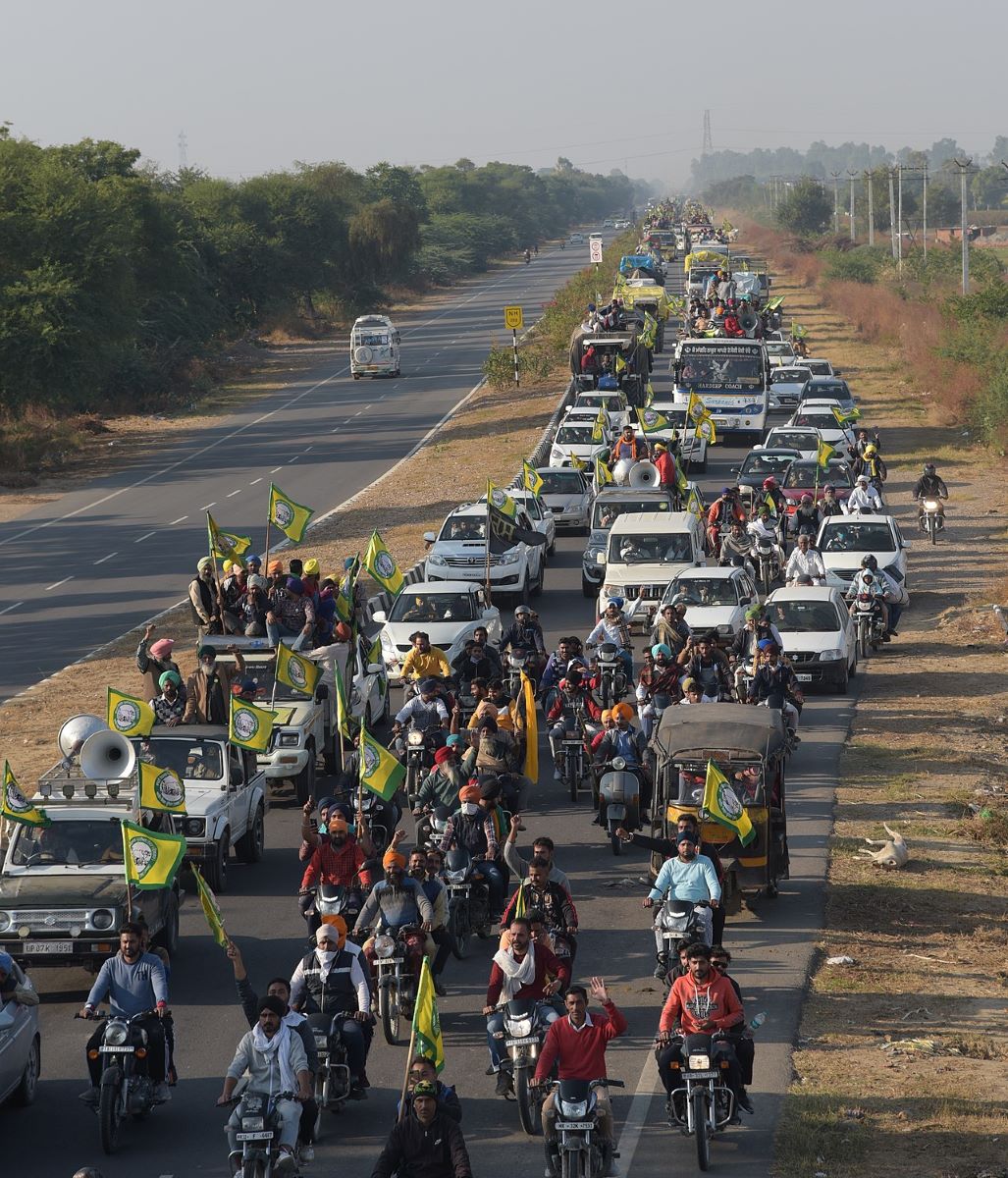A convoy of people with green and yellow flags occupies a lane of highway flanked by dry grass. People driving motorcycles are in the front, and behind them are rows of white cars and trucks.