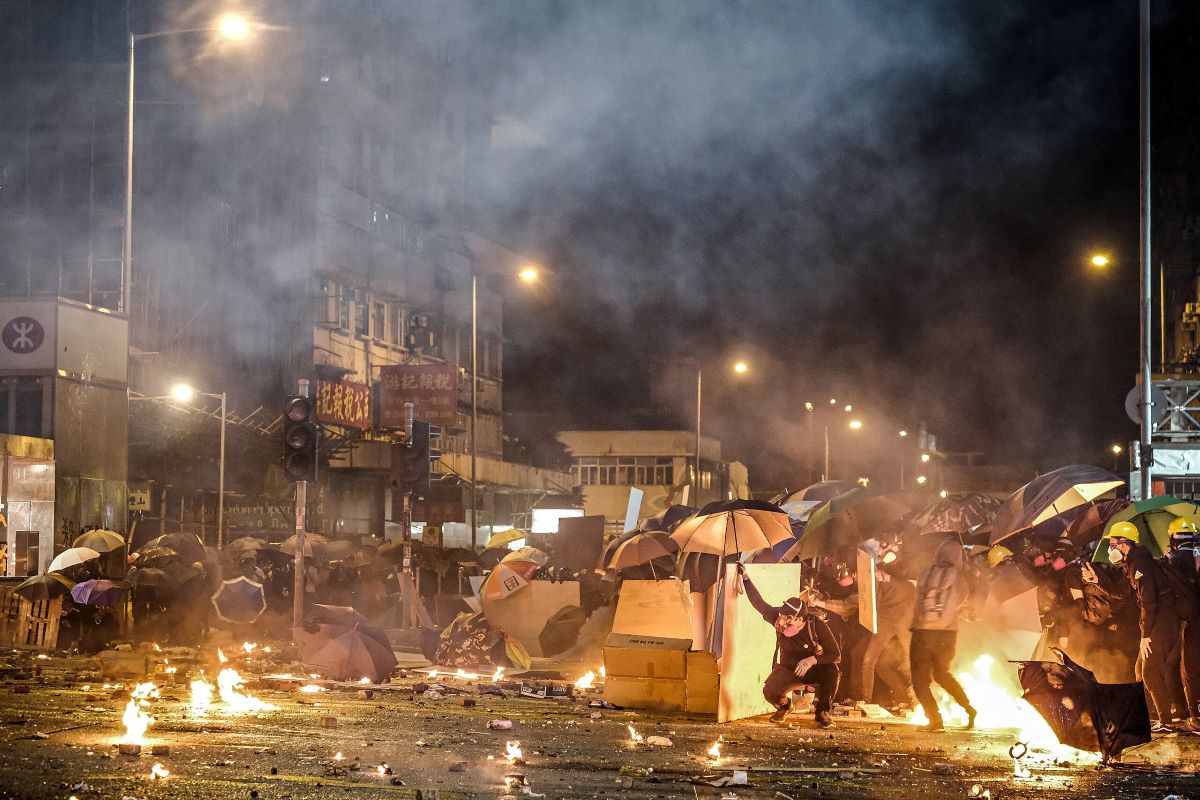 Several small items are in flames on the concrete before a makeshift cardboard barricade and many protesters holding umbrellas, wearing construction helmets and respirators. Grey smoke rises against the dark night sky. 