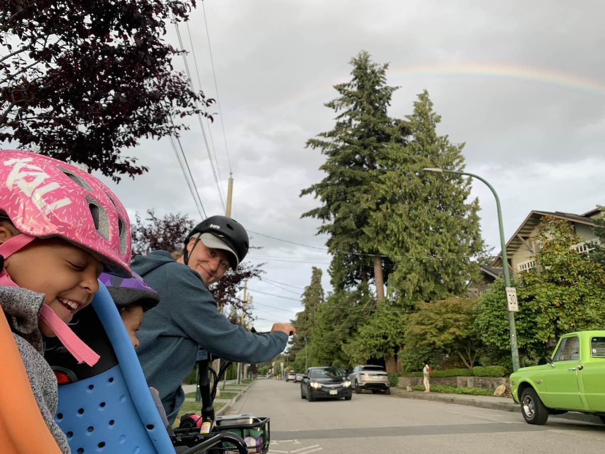 To the left of the frame, a woman in a grey-blue hoodie and bike helmet helms an e-bike with two young children in tow. They are riding down a quiet residential street on a grey day.