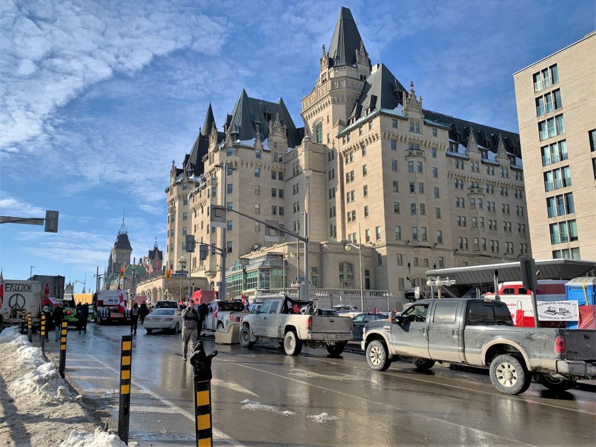 A line of large pick-up trucks snakes down a street lined with dirty snow in downtown Ottawa, where a large heritage building looms above the vehicles against a blue sky.