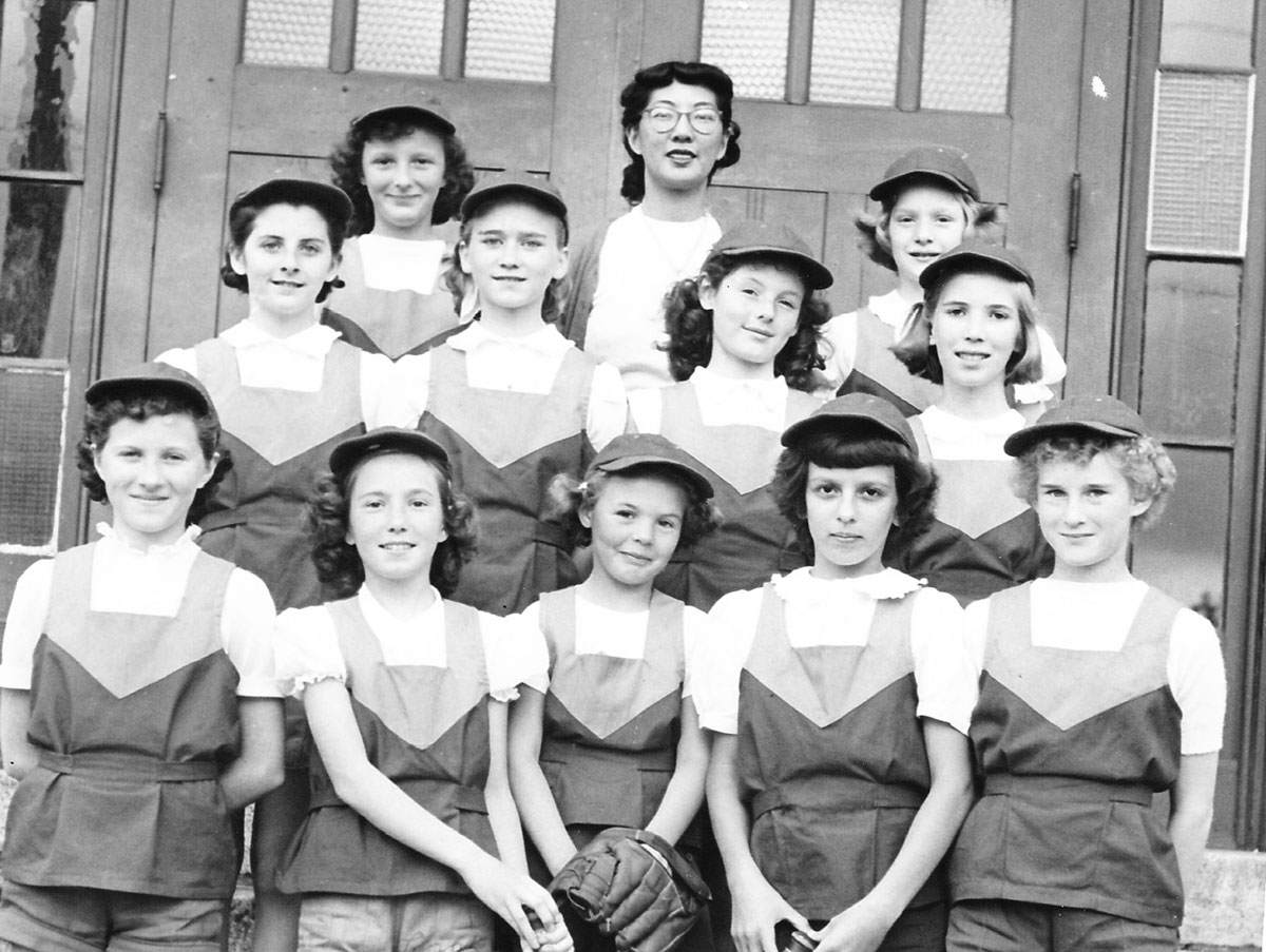 A black and white photo depicts Vivian Jung standing behind a group of elementary-school-aged girls in softball uniforms. Jung stands at the back of the group. She is wearing glasses and has her hair tied back.