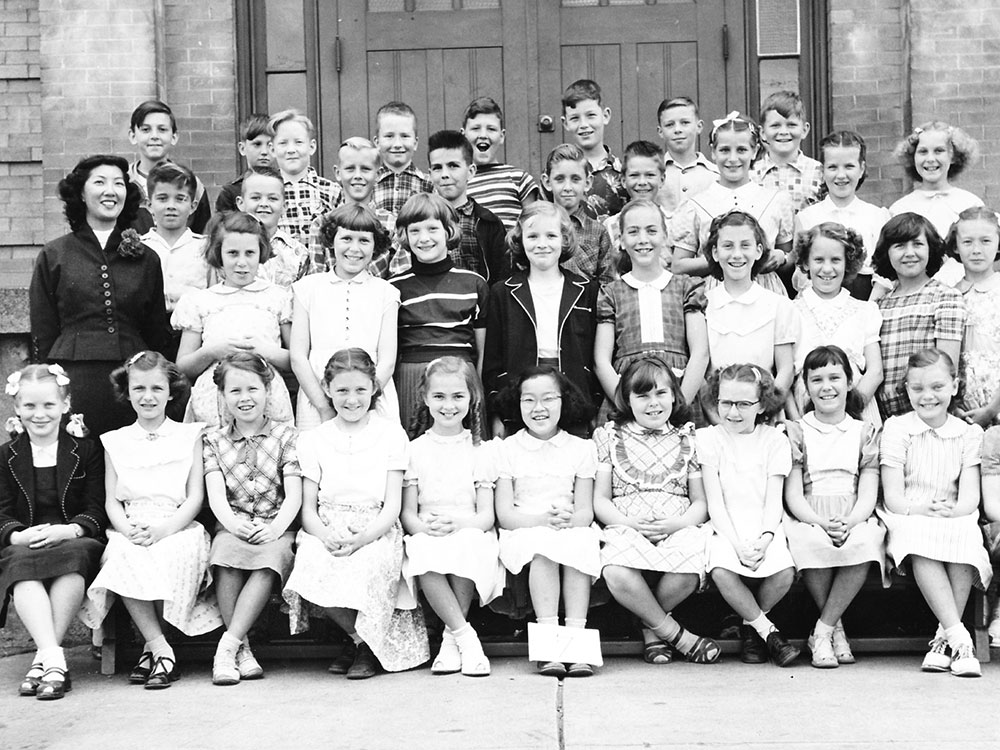 A black-and-white archival photo depicts Vivian Jung, to the left of the frame in a dark jacket, with the students of her Grade 4 class in 1953. Jung has wavy shoulder-length hair and is smiling. She is one of the only racialized people in the photo. 